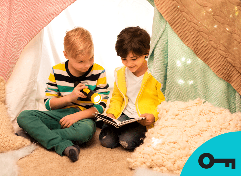 Deux enfant lisent un livre avec une lampe de poche dans une cabane de couvertures et de coussins.