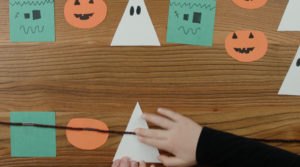 Un enfant fabrique une guirlande d'Halloween avec des carrés verts, des ovales orange et des triangles blancs